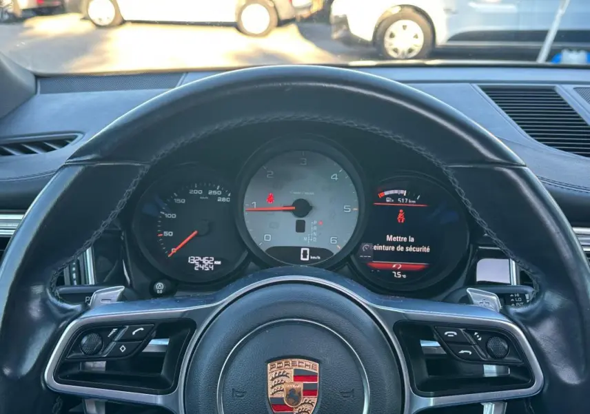 Vue rapprochée du tableau de bord et volant cuir noir du Porsche Macan gris foncé, affichant le compteur et alertes.