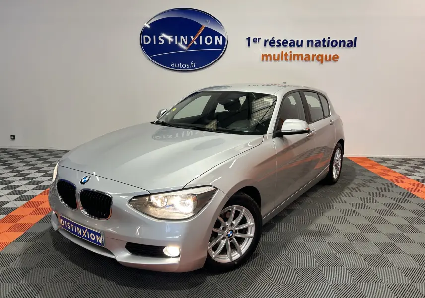 BMW Série 1 gris vue 3/4 avant droit avec phares allumés dans un showroom Distinxion.