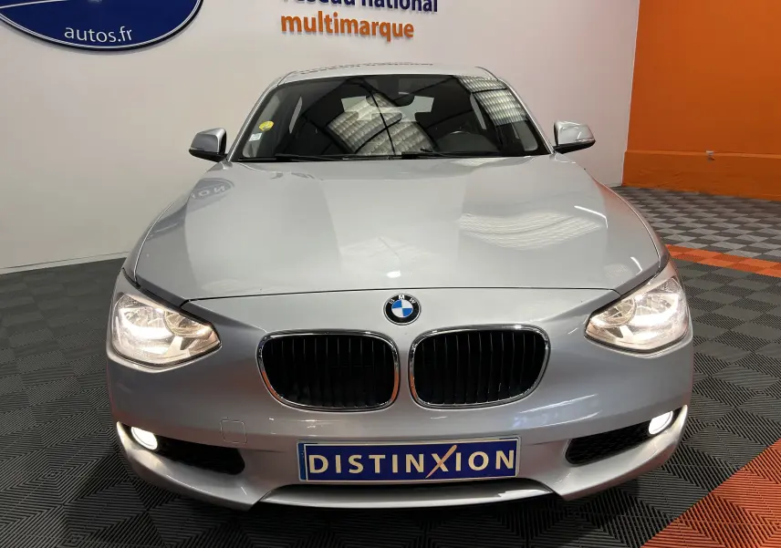 Vue frontale d'une BMW Série 1 gris clair avec phares allumés et plaque Distinxion en concession multimarque.