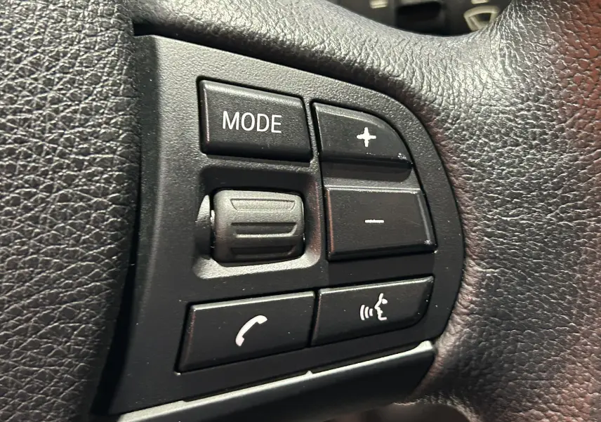 Gros plan sur les commandes noires du volant en cuir de la BMW Série 1 gris diesel, avec boutons mode et volume.