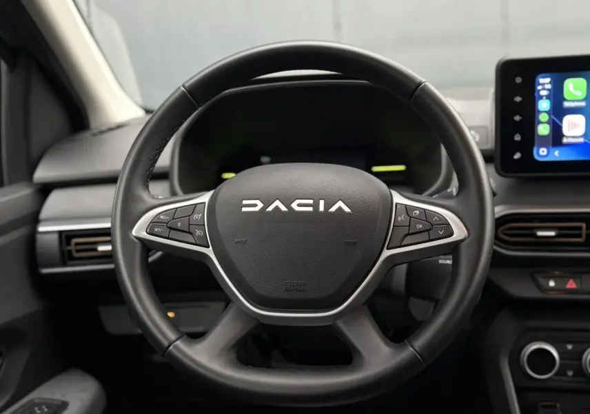 Vue rapprochée du volant noir DACIA avec commandes intégrées, tableau de bord et écran tactile à droite visibles.