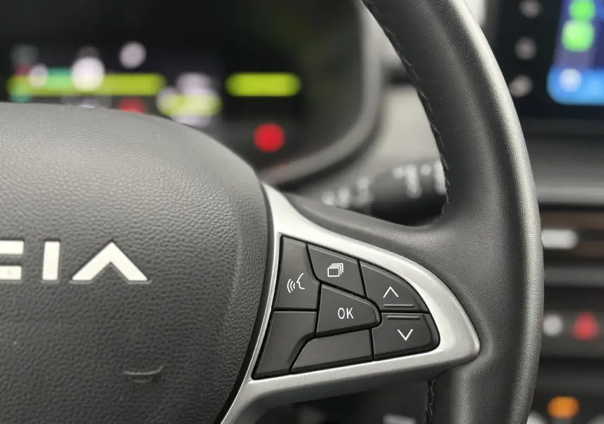 Gros plan sur le volant côté droit du Dacia Jogger gris urbain avec commandes multifonctions et tableau de bord flou.