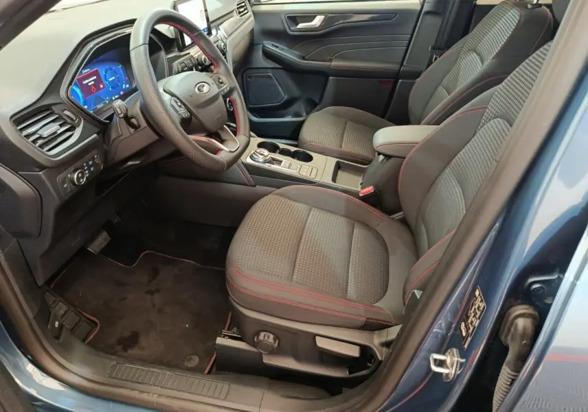 Vue intérieure côté conducteur du Ford Kuga bleu 2024, avec sièges tissu gris à surpiqûres rouges et tableau de bord digital.