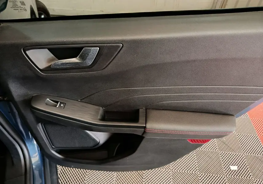Vue intérieure côté gauche de la porte avant d’un Ford Kuga bleu, avec poignée argentée et accoudoir noir à surpiqûres rouges.