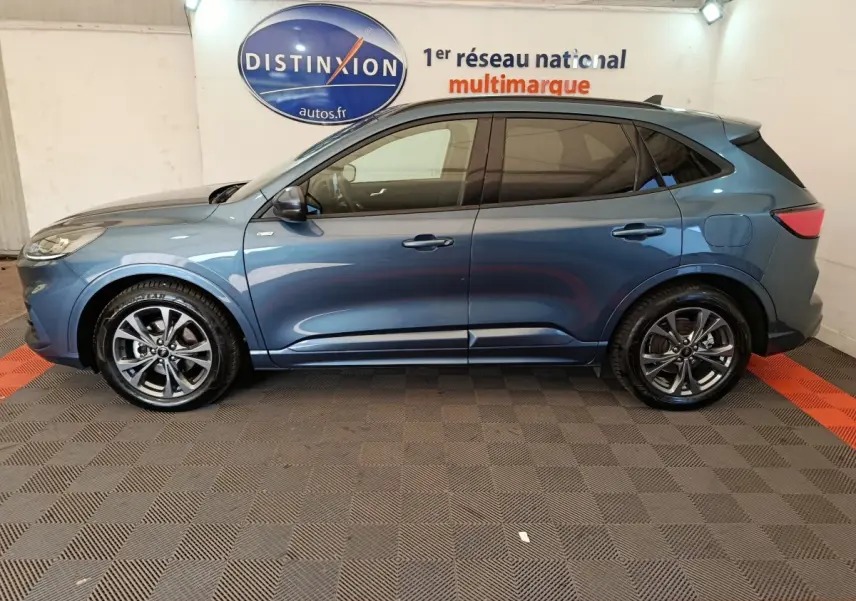 Vue latérale droite d’un Ford Kuga 2024 bleu métallisé St Line avec jantes alliage et vitres teintées, en intérieur showroom.