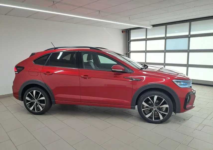 Vue de profil côté gauche d'un Volkswagen Taigo Rouge Roi avec jantes alliage noires brillantes et toit panoramique en intérieur showroom.