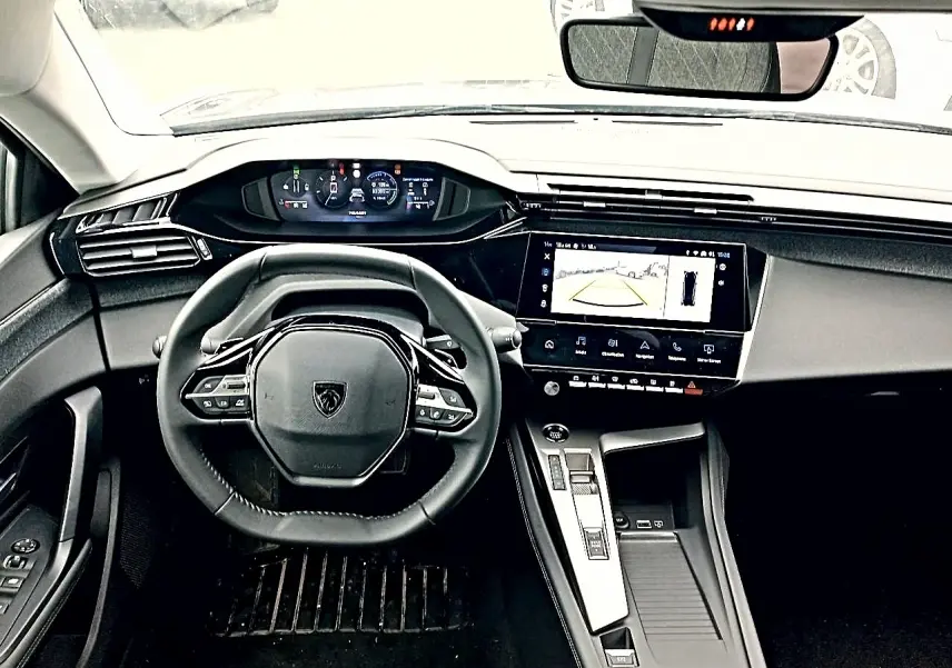 Vue intérieure frontale du tableau de bord et volant cuir noir de la Peugeot 408 Hybrid 145 allure 2024 avec écran tactile et caméra de recul affichée.
