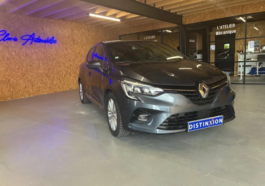 Renault Clio gris 3/4 avant droit dans un showroom avec éclairage intérieur et logo bleu au mur.