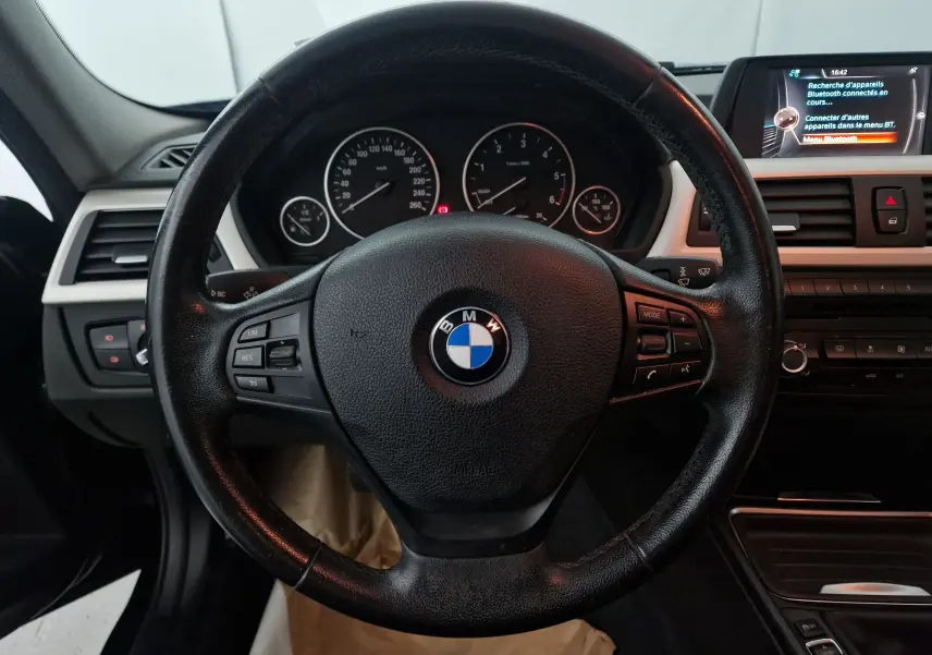 Vue rapprochée du volant noir d'une BMW Série 3 316D 2014 avec tableau de bord et écran multimédia visibles.