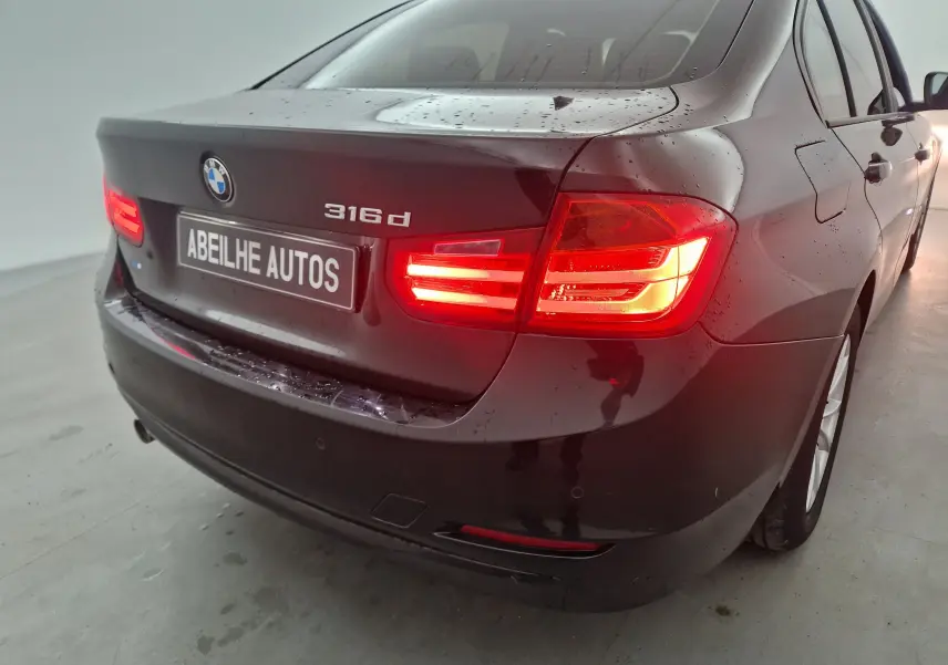 Vue 3/4 arrière droite d'une BMW Série 3 316d noire avec feux arrière allumés et carrosserie mouillée.