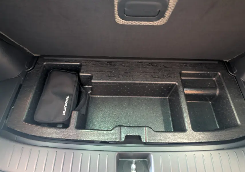 Coffre ouvert du Kia Sportage blanc 2025, vue plongeante sur compartiment de rangement avec trousse de mobilité noire.