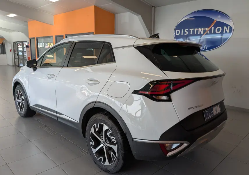 KIA Sportage blanc vue 3/4 arrière droit en intérieur, avec jantes alliage 18’’ et barres de toit visibles.