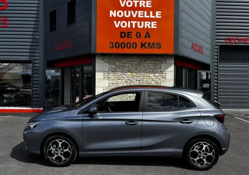 Vue latérale gauche d'une MG3 gris clair 2025 garée devant un bâtiment avec panneau orange "Votre nouvelle voiture".