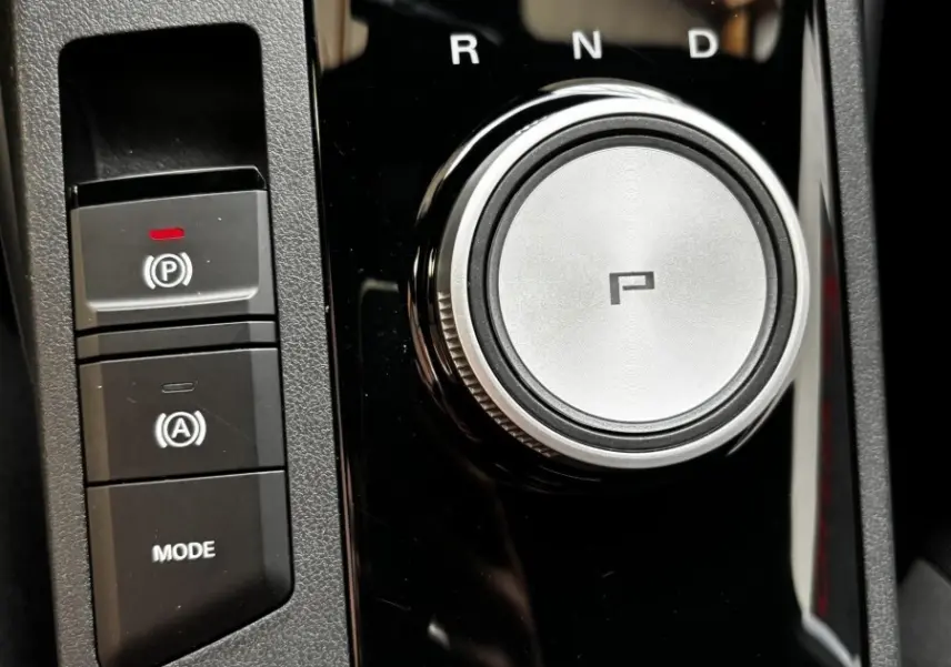 Vue rapprochée du sélecteur rotatif de boîte automatique et des boutons de frein à main et mode sur console noire brillante d’une MG3 gris clair.