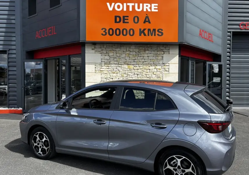 Vue latérale droite d'une MG3 gris clair garée devant un bâtiment avec panneau orange "Votre nouvelle voiture de 0 à 30000 kms".