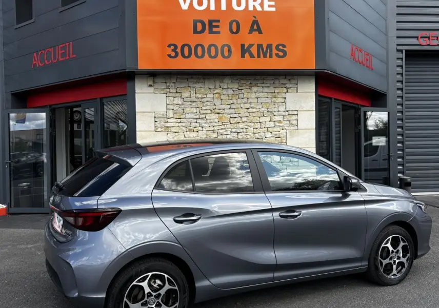 Vue latérale droite d'une MG3 gris clair 2025 garée devant un bâtiment avec panneau orange "Votre nouvelle voiture".