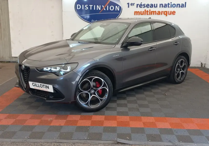 Alfa Romeo Stelvio gris foncé en 3/4 avant droit, avec jantes distinctives et phares LED allumés.
