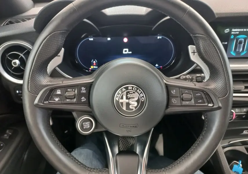 Vue rapprochée du volant cuir noir d'une Alfa Romeo Stelvio 2024 avec tableau de bord numérique et bouton start/stop visible.