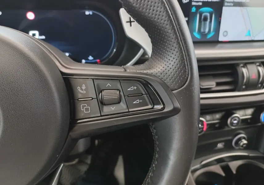 Gros plan sur le volant cuir perforé côté gauche de l'Alfa Romeo Stelvio 2024 avec commandes multifonctions et écran tactile en arrière-plan.