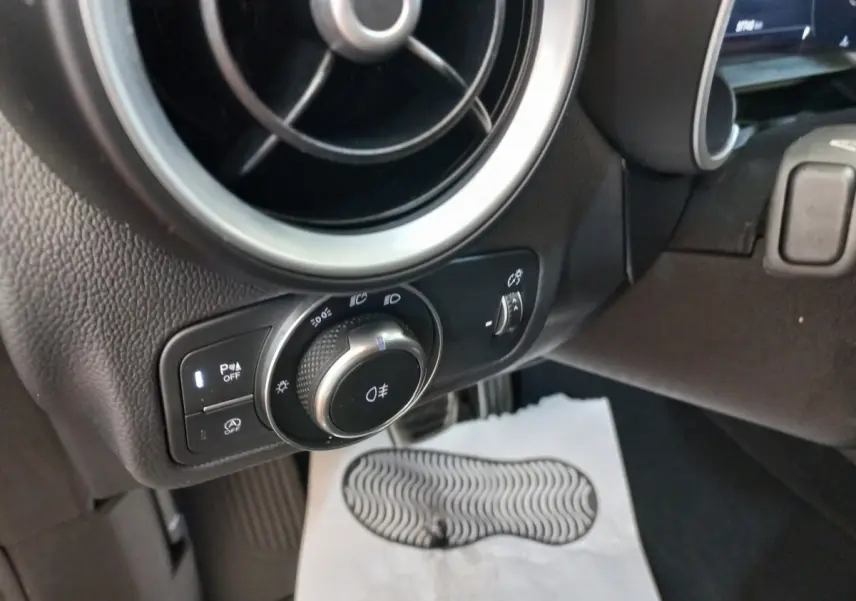 Gros plan sur la commande des phares et l'aération ronde côté gauche du tableau de bord gris foncé de l'Alfa Romeo Stelvio.