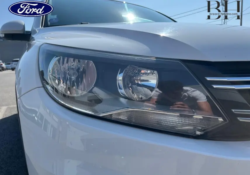 Gros plan sur le phare avant gauche d'un Volkswagen Tiguan blanc Candy, avec calandre noire et chrome visible.