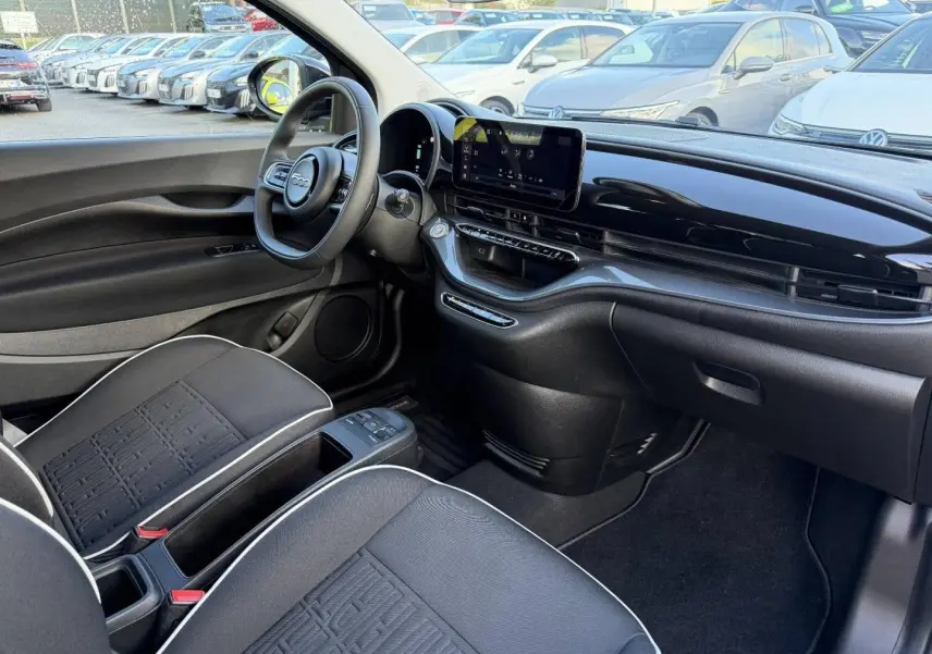 Vue intérieure côté conducteur de la FIAT 500 e 118ch MY24 avec tableau de bord noir brillant et sièges tissu noir monogramme.