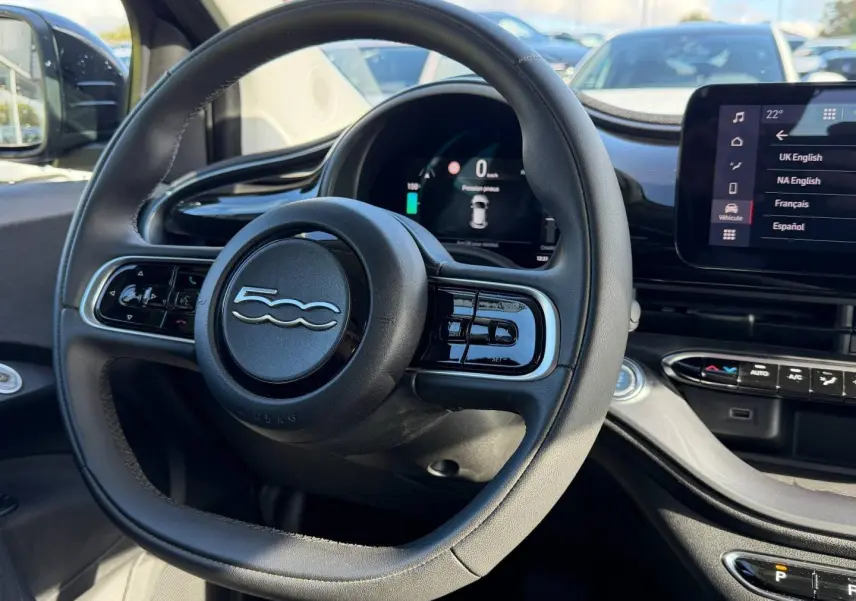 Vue rapprochée du volant noir multifonction avec logo 500 et tableau de bord numérique de la Fiat 500 e 118ch MY24.