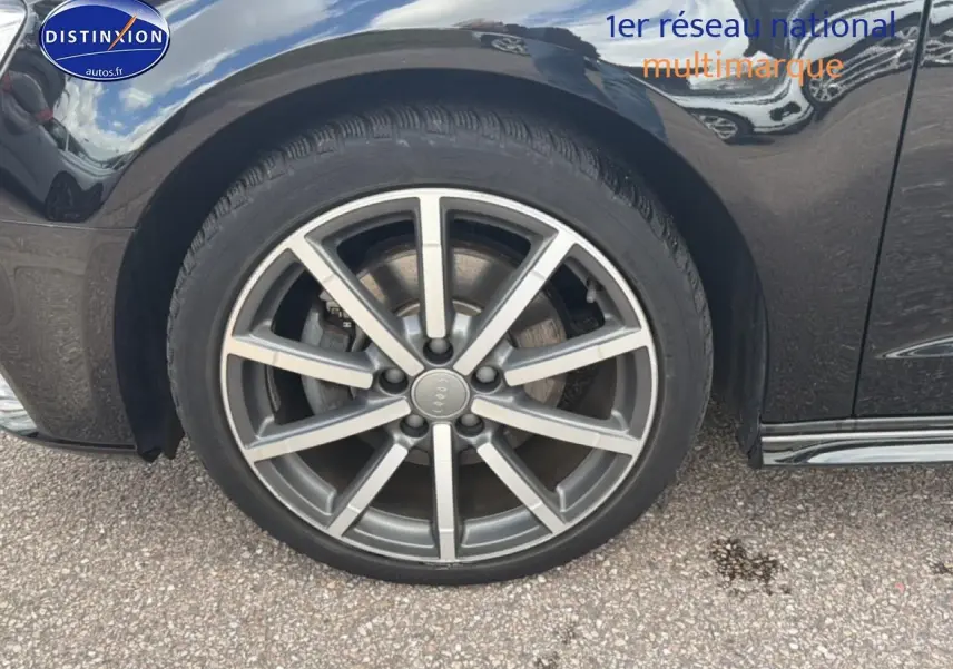 Gros plan sur la roue avant gauche en jantes alu d'une Audi A3 Sportback noire, avec reflet du ciel sur la carrosserie.