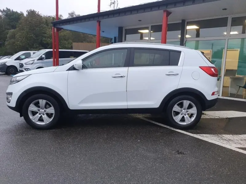 Profil côté gauche d'un KIA SPORTAGE blanc de 2013, avec barres de toit et jantes alliage visibles.