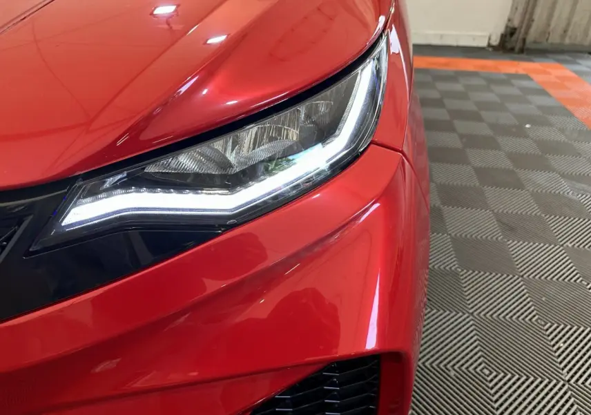 Gros plan sur le phare avant droit allumé d'une Aixam Ambition rouge, avec une partie du pare-chocs visible.