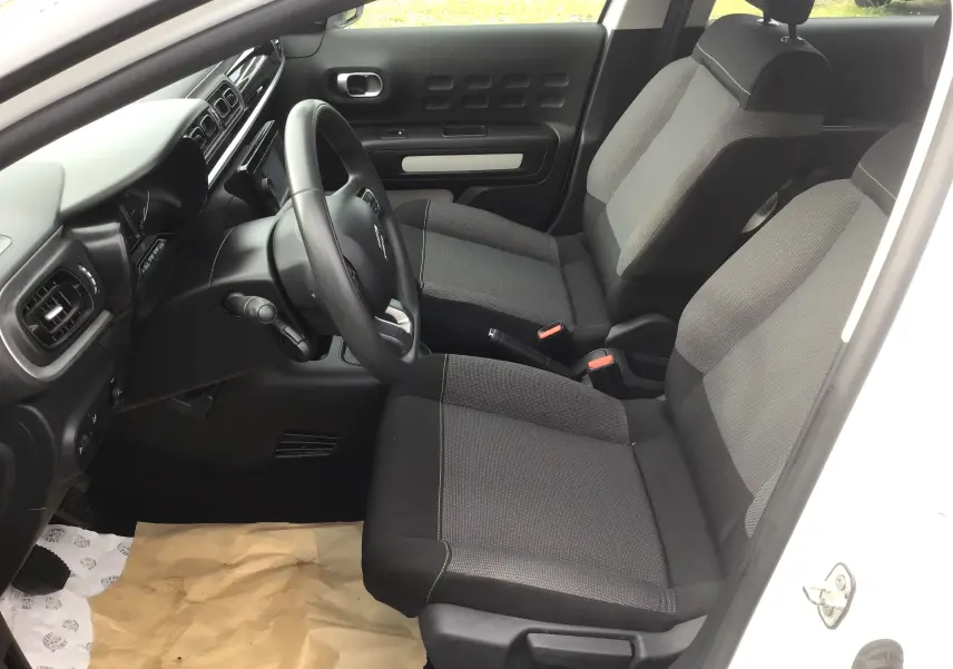 Vue intérieure côté conducteur de la Citroën C3 blanche 2021, sièges tissu gris avec surpiqûres jaunes et tableau de bord noir.