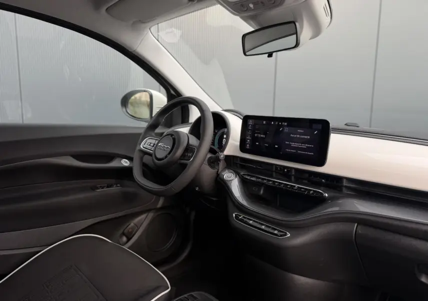 Vue intérieure côté conducteur de la Fiat 500 électrique 2024, tableau de bord blanc avec écran tactile et volant noir.