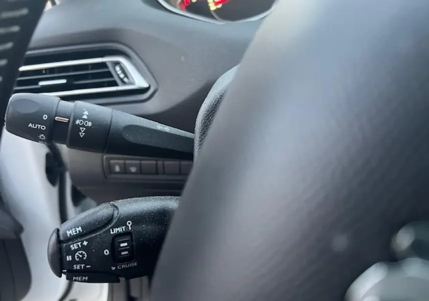 Gros plan sur les commandes au volant côté gauche d'une Peugeot 308 blanche, mettant en valeur les fonctions régulateur et éclairage.