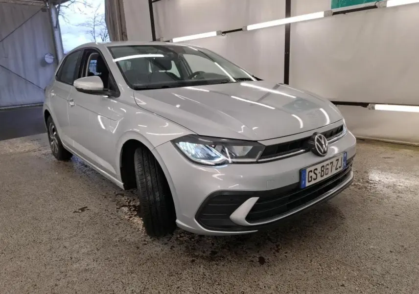 Volkswagen Polo gris clair vue 3/4 avant droit avec phares allumés dans un garage intérieur.
