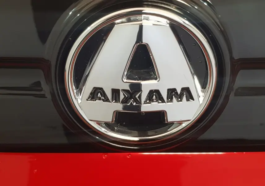 Gros plan sur le logo chromé AIXAM sur fond noir et rouge vif de la carrosserie d'une Aixam Ambition 2026.