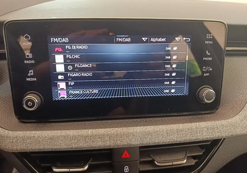 Écran tactile central de la Skoda Scala 2024 affichant les stations radio FM/DAB, entouré de commandes et finition tissu grise.