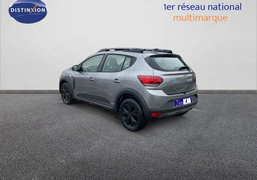 Vue 3/4 arrière droite d'une Dacia Sandero Stepway gris schiste métal avec jantes noires et barres de toit noires.