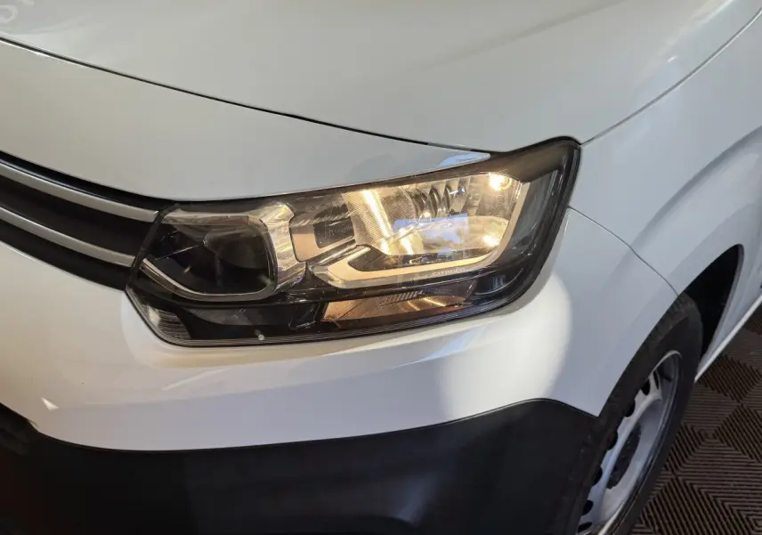 Gros plan sur le phare avant droit allumé d’un Citroën Berlingo blanc avec calandre noire et jante acier.