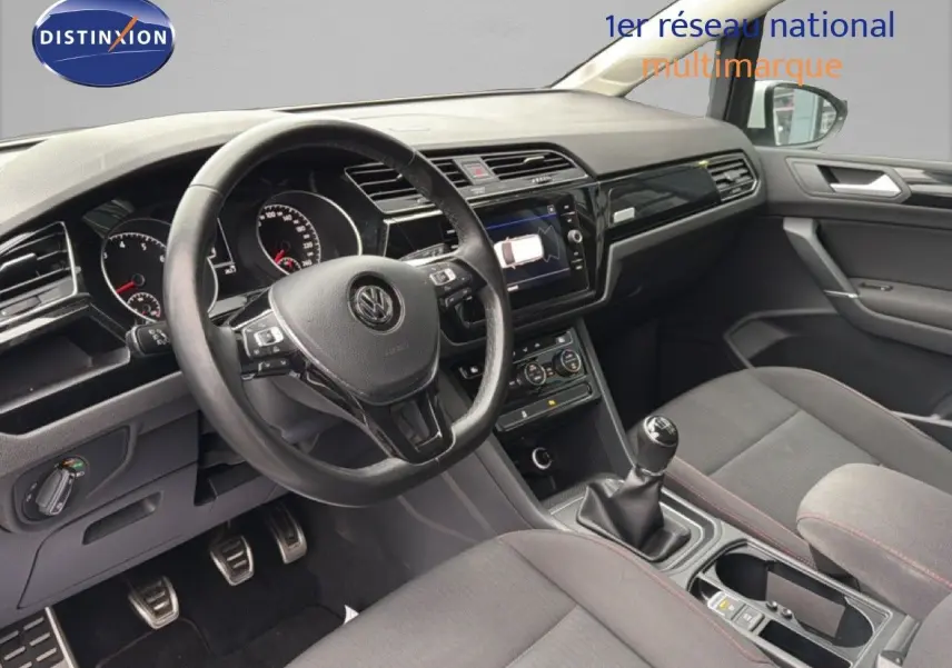 Vue intérieure côté conducteur du Volkswagen Touran 2018, montrant le volant, la console centrale et les sièges tissu gris.