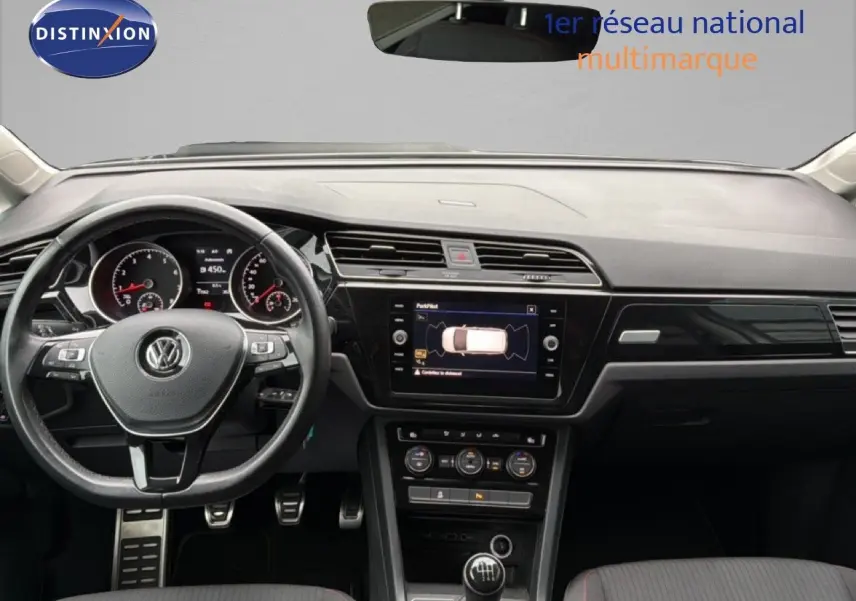 Vue intérieure frontale du tableau de bord et volant noir d’un Volkswagen Touran blanc avec boîte manuelle et écran central tactile.