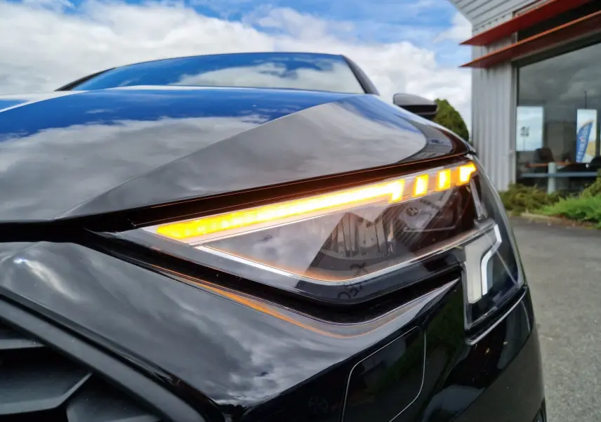 Gros plan sur le feu avant droit allumé d'une Audi A3 Sportback noire, soulignant ses lignes modernes et précises.