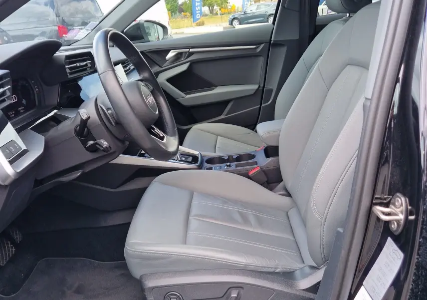 Vue intérieure côté conducteur de l'Audi A3 Sportback noire, mettant en valeur les sièges en cuir gris et le tableau de bord moderne.