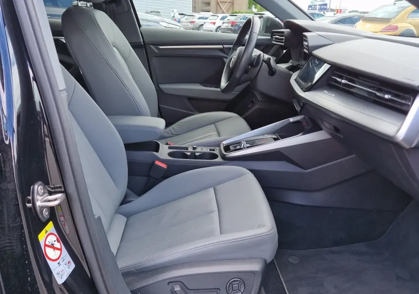 Vue intérieure côté conducteur de l'Audi A3 Sportback noire, mettant en valeur les sièges en cuir gris et le tableau de bord moderne.