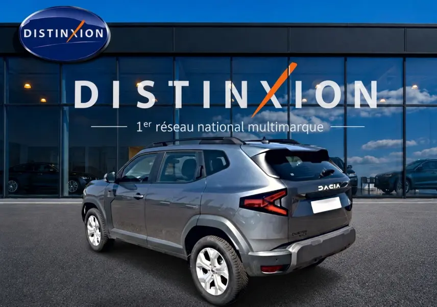 Vue 3/4 arrière droite d'un Dacia Duster gris schiste 2026 avec barres de toit et jantes alu devant un showroom Distinxion.