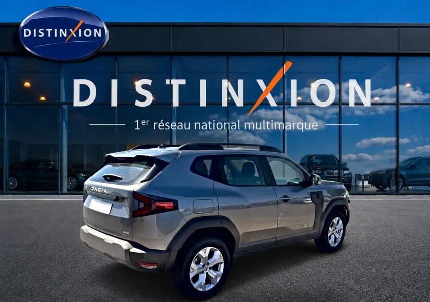 Dacia Duster gris schiste en 3/4 arrière droit, avec barres de toit et jantes alu visibles devant un bâtiment Distinxion.