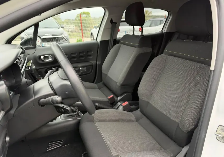 Vue intérieure côté conducteur d'une Citroën C3 blanche, montrant les sièges tissu gris et la console centrale.