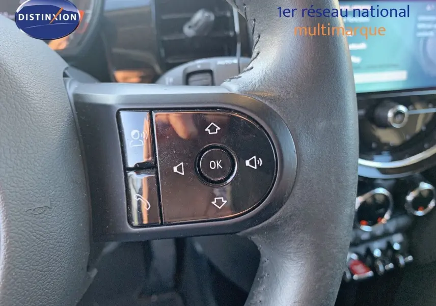 Gros plan sur les commandes noires du volant d'une MINI 1.5 136CH rouge chili, avec tableau de bord flou en arrière-plan.