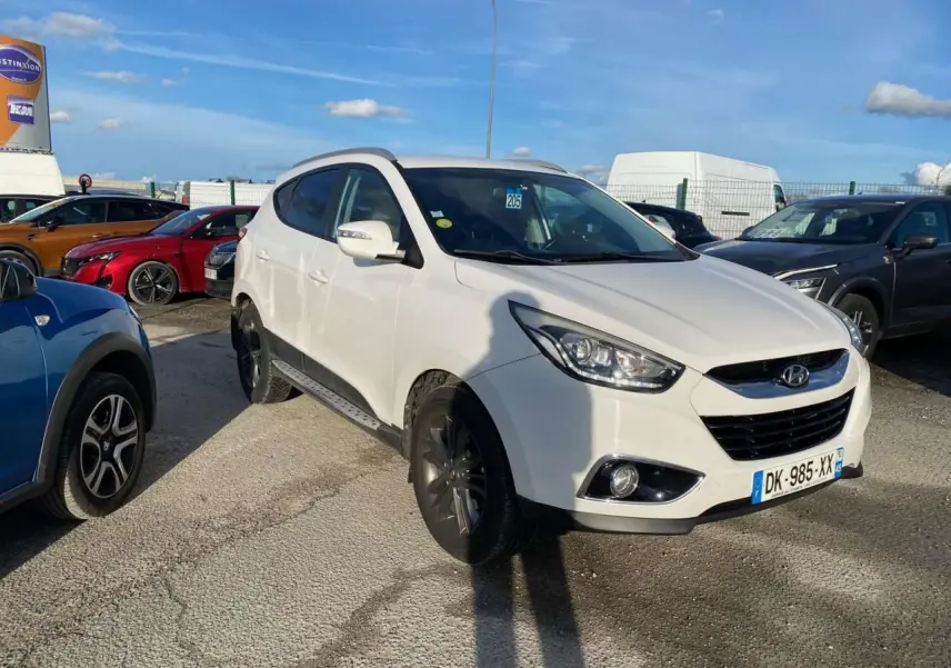Hyundai ix35 blanc vu en 3/4 avant droit sur un parking, avec jantes foncées et projecteurs allumés.
