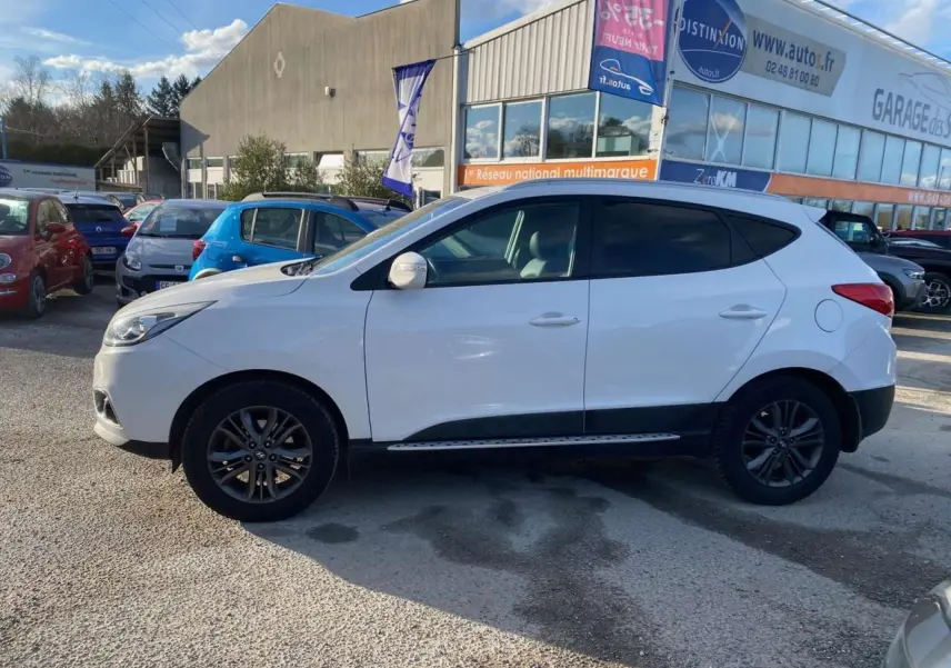 Vue de profil côté gauche d'un Hyundai ix35 blanc stationné devant un garage, avec jantes alliage foncé.