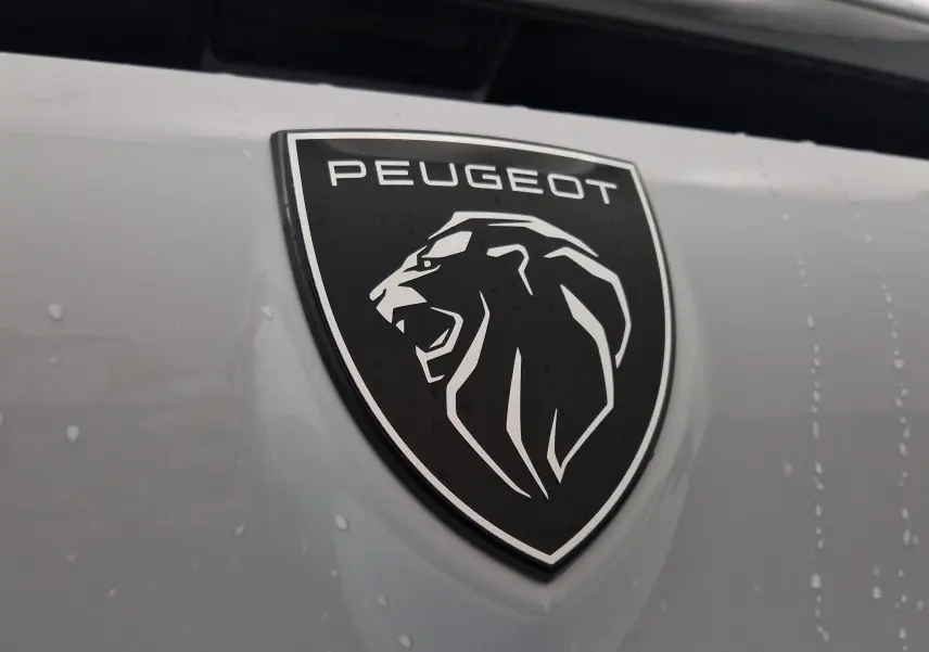 Gros plan sur l'emblème noir et blanc du lion Peugeot sur la calandre blanche Okenite d'une 308 2025.